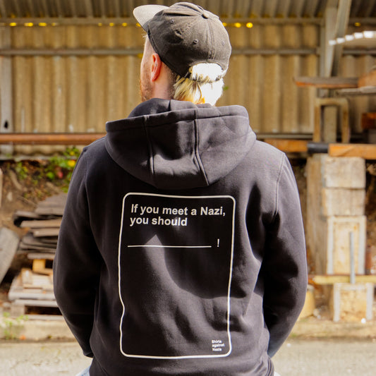 Männliches Modell mit Cap, von hinten zu sehen. Das Modell trägt einen schwarzen Hoodie mit einem weißen, handgemachten Aufdruck. Der Aufdruck ließt "If you meet a Nazi you should?" und erinnert an eine Spielkarte.