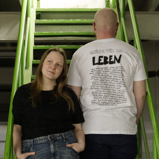 Zwei Models auf grüner Treppe. Sie trägt ein schwarzes Shirt ohne Frontprint. Er trägt ein weißes Shirt mit Rückenprint. "Gründe für das Leben" handgedruckt mit schwarzer Farbe auf weißes Shirt.