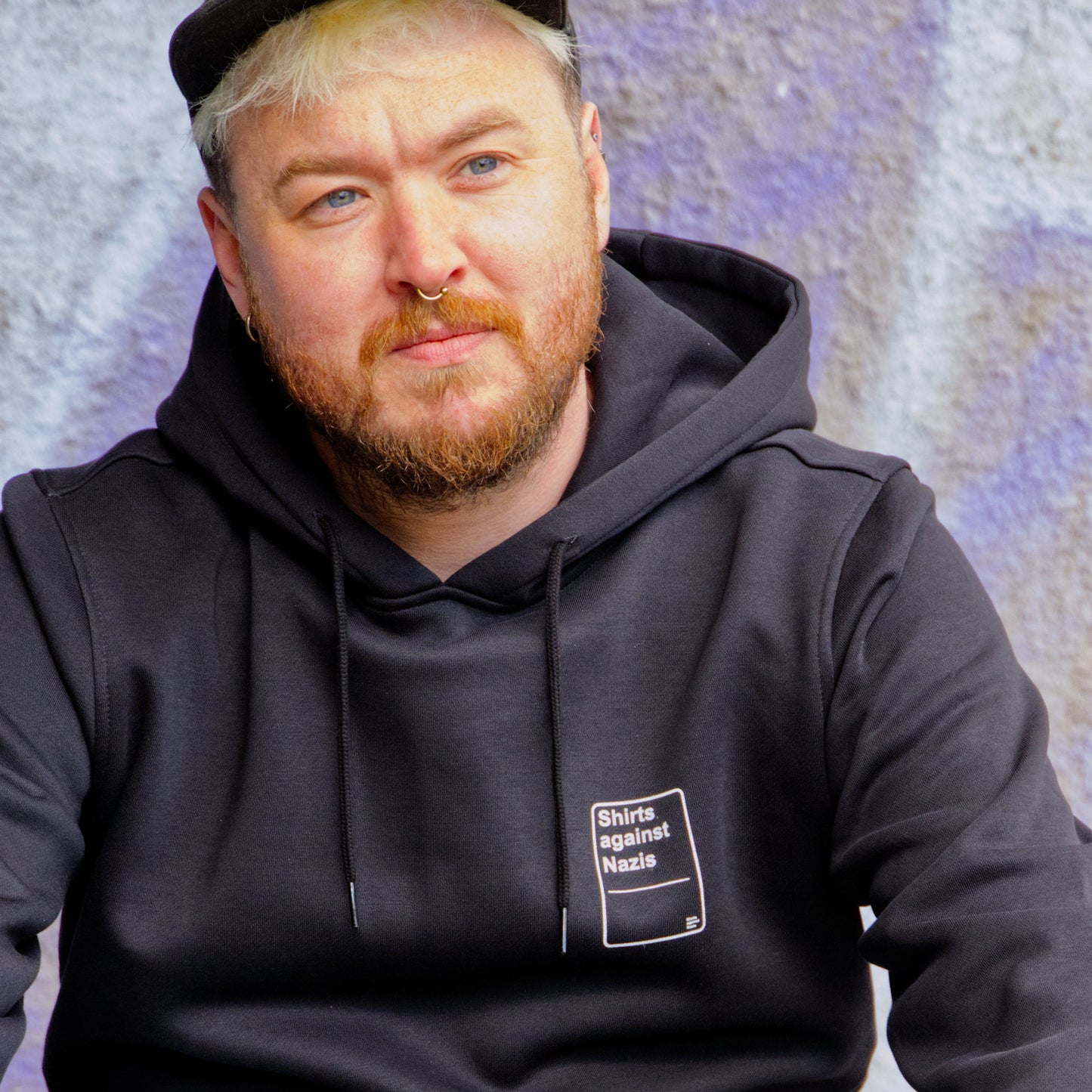 Männliches Modell mit Cap, vor lila Hintergrund. Das Modell trägt einen schwarzen Hoodie mit einem weißen, handgemachten Aufdruck. Der Aufdruck ließt " Shirts against Nazis und erinnert an eine Spielkarte.