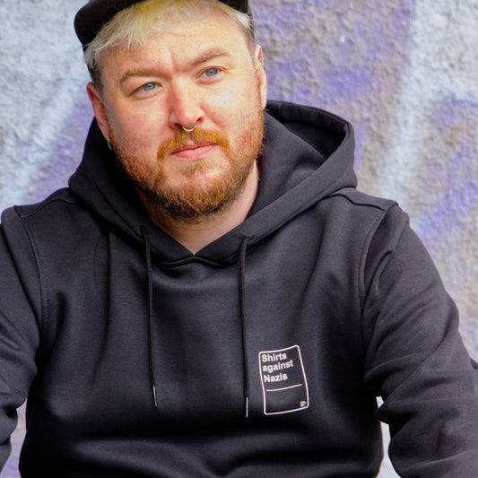 Männliches Modell mit Cap, vor lila Hintergrund. Das Modell trägt einen schwarzen Hoodie mit einem weißen, handgemachten Aufdruck. Der Aufdruck ließt " Shirts against Nazis und erinnert an eine Spielkarte.