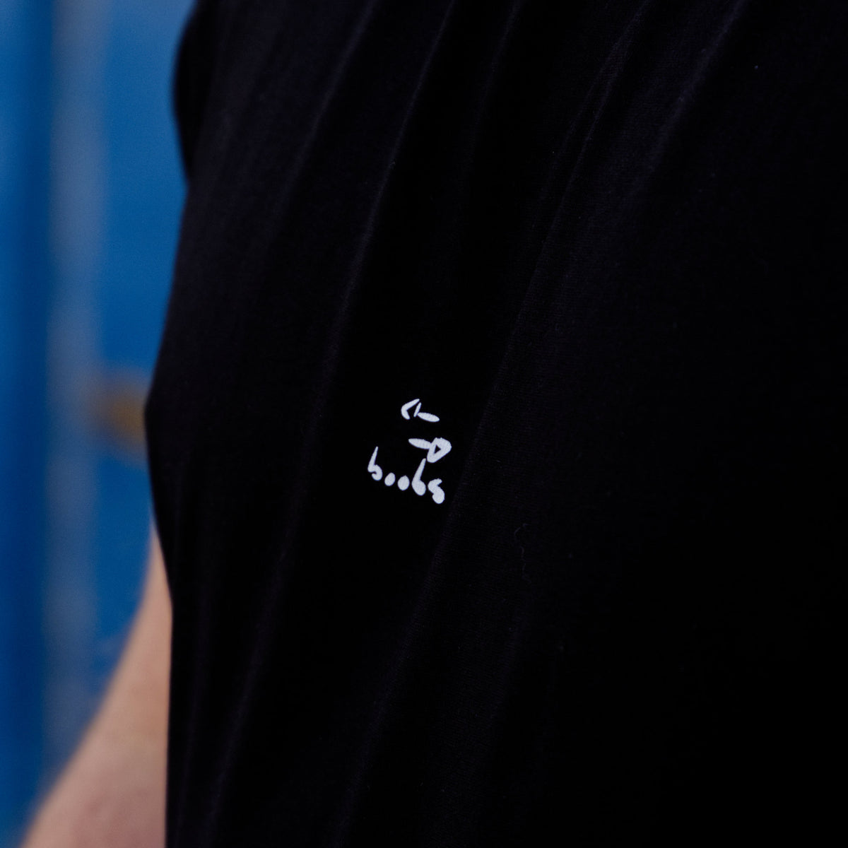 Männliches Modell mit schwarzem Shirt vor blauem Hintergrund. Auf das Shirt ist das Motiv "boobs" aufgedruckt.